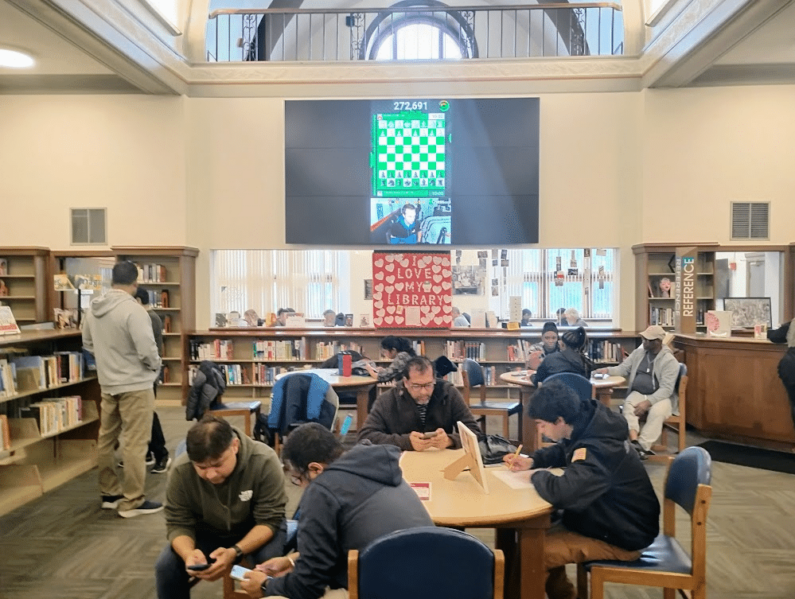 An online chess player plays chess on a digital screen, behind the windows in a study room a local chess tournament plays. Community members sit at tables doing homework and making conversation. [All photos by Kelly Margaret Heikkla]